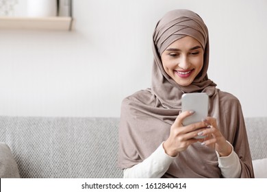 Portrait of millennial muslim girl in hijab using smartphone at home, messaging or browsing social networks, sitting on sofa in living room