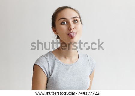 Portrait of mischievous spoiled teenage girl with hair bun fooling around, looking at camera and sticking out her tongue as if teasing at someone. Human facial expressions and body language