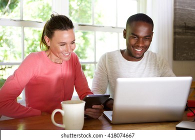 Portrait Of Mixed Race Couple Sitting At Table Using Laptop And Digital Tablet Computer At Home