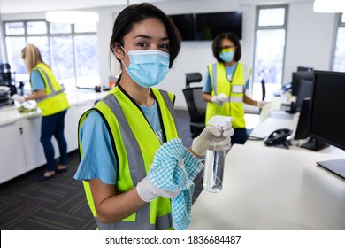 Portrait of mixed race woman wearing hi vis vest, gloves and face mask sanitizing office with disinfectant, colleagues in the background. Hygiene in workplace during Coronavirus Covid 19 pandemic. - Powered by Shutterstock