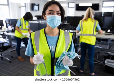 Portrait Of Mixed Race Woman Wearing Hi Vis Vest, Gloves And Face Mask Sanitizing Office With Disinfectant, Colleagues In The Background. Hygiene In Workplace During Coronavirus Covid 19 Pandemic.