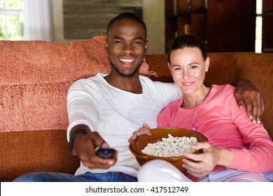 Portrait Of Mixed Race Young Couple Sitting On Living Room Sofa Watching A Movie And Eating Popcorn
