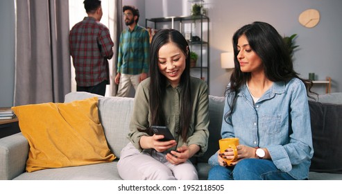 Portrait Of Mixed-race Female Friends Sitting Together At Home On Sofa And Chatting Typing On Smartphone Gadget While Asian And Hindu Young Guys Speaking On Background. Leisure Concept