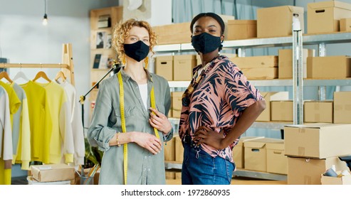 Portrait of mixed-race young females standing in clothing shop warehouse wearing masks on faces and looking at camera. Clothing designer. Sale assistant in storehouse. Fashion industry concept - Powered by Shutterstock