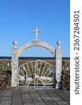portrait mode photo of the entrance to a cemetery in iceland with a white stone arch with a cross, a white gate and low volcanic stone walls on the sides, the sea in the background and a blue sky