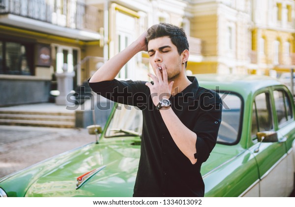 Portrait of a model young\
handsome sexy male brunette guy with dark skin Turkish Middle\
Eastern brunette posing smoking a cigarette near old retro car on\
the street.