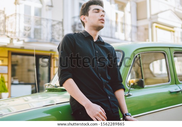 Portrait of a model young\
handsome sexy male brunette guy with dark skin Turkish Middle\
Eastern brunette posing smoking a cigarette near old retro car on\
the street.