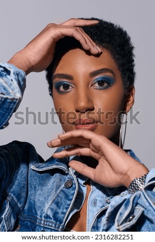 Portrait of modern african american woman with short hair and bold makeup touching face and looking at camera while posing in denim outfit isolated on grey, denim fashion concept
