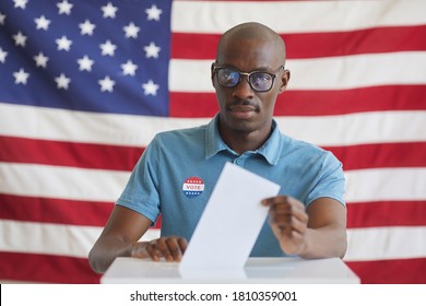 Portrait of modern African-American man putting vote bulletin in ballot box and looking at camera while standing against American flag on election day, copy space - Powered by Shutterstock