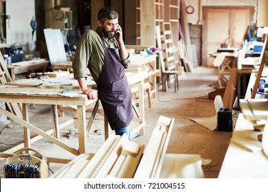 Portrait of modern bearded carpenter speaking by phone in woodworking shop taking break from work