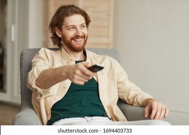 Portrait of modern bearded man watching TV at home while relaxing in comfy armchair and holding remote, copy space