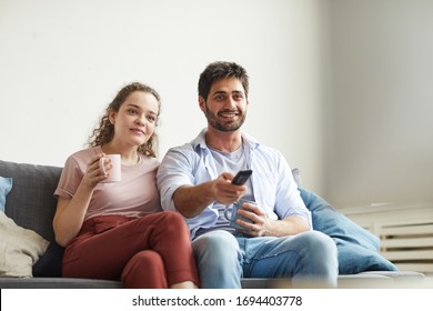 Portrait of modern couple watching TV and holding mugs while sitting on sofa at home in cozy apartment enjoying lazy time, copy space