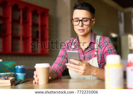 Portrait of modern creative woman using smartphone and drinking coffee while taking break from work in art studio