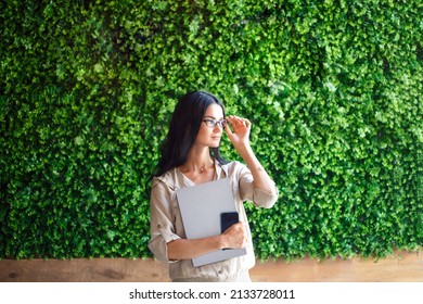 Portrait modern female woman using laptop and phone, communicates on internet with customer in city cafe. Cozy office workplace. Eco-friendly background wall of green grass - Powered by Shutterstock