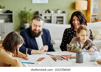 Portrait Of Modern Jewish Family Drawing Together While Sitting At Table , Focus On Smiling Father Wearing Kippah