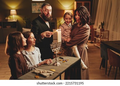 Retrato de moderna jewish familia iluminando velas menorah plateadas durante la celebración de Hanukkah en un acogedor hogar