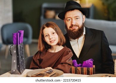 Portrait Of Modern Jewish Man With Daughter Looking At Camera While Sitting At Dinner Table In Cozy Home Setting