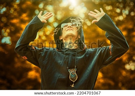 Portrait of a modern male shaman in black ethnic cloak with dreadlocks, tattoos and sunglasses  turning to heaven and performing a ritual outdoors. Shamanic rituals. 