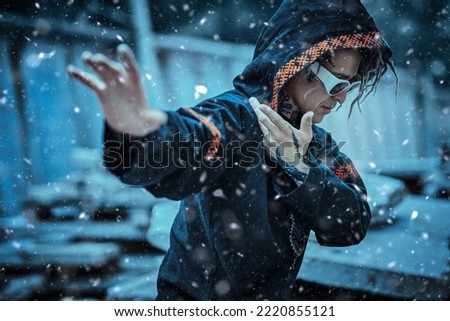 Portrait of a modern male shaman in black ethnic cloak with dreadlocks, tattoos and sunglasses performing a ritual on the street on a snowy day. Shamanic rituals. 