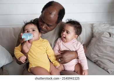 Portrait of modern mature Black man sitting on sofa taking care of his twin babies starting feeding them with formula