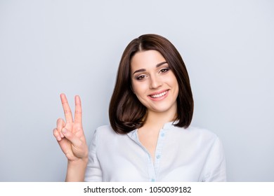 Portrait of modern, smiling, nice girl with short hairstyle showing v-sign, two fingers, looking to the camera, isolated on grey background