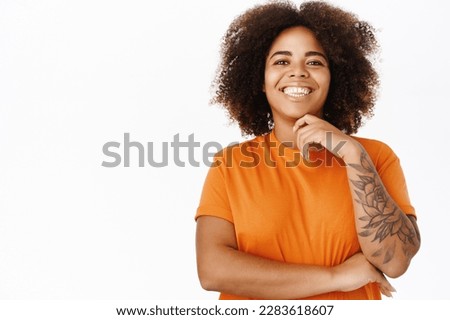 Portrait of modern smiling woman with tattoos, wears orange t-shirt, looks happy, stands over white background.