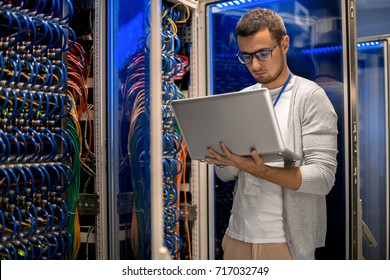 Portrait of modern young man holding laptop standing in server room working with supercomputer in blue light