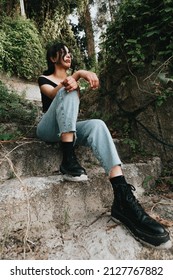 Portrait Of A Modern Young Woman Sitting On Stairs With Military Boots And Mom Jeans. Styling And Fashion Concepts. Free Time On Young People And Leisure Time. Travel On Vacation