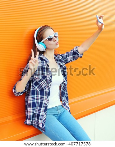 Portrait of modern young woman taking selfie with smartphone in wireless headphones listening to music blowing lips sending sweet air kiss on orange background