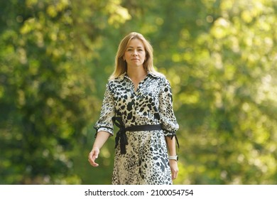 Portrait Of The Modern Young Woman Walking At The Moscow Public Park. Blonde With Long Hair. Stern Look. Defocused Trees With Lush Foliage As Background. Lifestyle Concept. Sunny Summer Day.