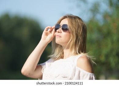 Portrait Of The Modern Young Woman Walking At The Natural Parkland. Blonde With Long Hair. Stern Look. Defocused Trees With Lush Foliage As Background. Lifestyle Concept. Sunny Summer Day.