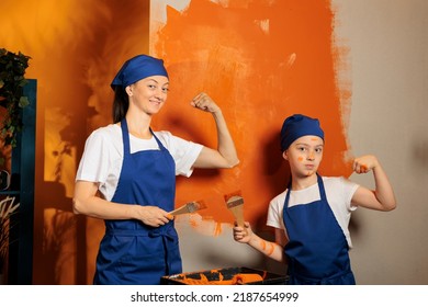 Portrait Of Mom And Kid Flexing Arm Muscles While Painting Room Walls With Orange Color Paint And Brush. Woman With Little Child Using Renovating Tools, Working Together On House Decor.