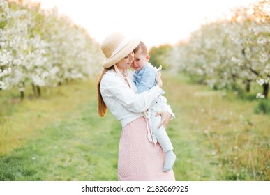 Portrait Of Mom In A Straw Hat With Crying Son In Her Arms In The Spring Garden