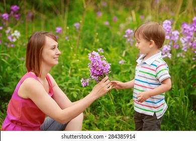 Portrait Of Mother And Son Together Sitting In On Flower Meadow Field  In Countryside Village, Smiling, Holding And Smelling Flowers, Happy Family Concept, Summer Fun Activity, Mothers Day Holiday