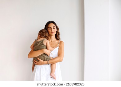 Portrait of motherhood. Young beautiful loving mother wearing white dress holding cute baby boy while standing isolated over white wall. Mom cuddling little son and expressing unconditional love - Shutterstock ID 2103982325