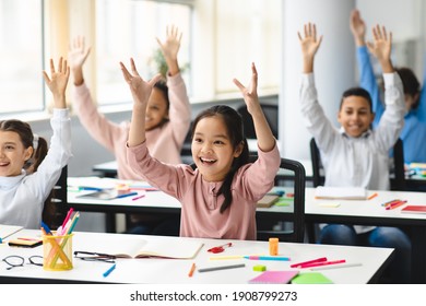 Portrait of multiciltural excited diverse group of small happy elementary school children sitting at desks in classroom, raising hands high up and waving, having fun, learning with pleasure
