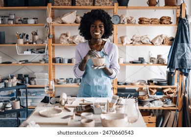 Portrait of multicultural female pottery class student standing at pottery workshop with earthenware in hands and smiling at the camera. Smiling young diverse craftswoman posing at ceramics studio - Powered by Shutterstock