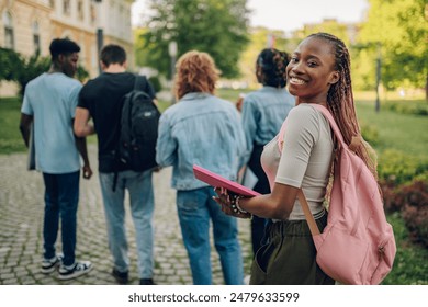 Portrait of multicultural female university student standing at campus near college building with textbook in hands and backpack and smiling at the camera. In background classmates rushing on lecture.
