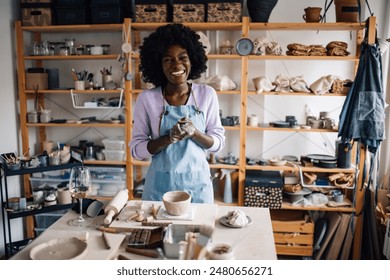 Portrait of multicultural pottery designer standing at ceramics studio surrounded by clay works and smiling at camera. Cheerful female potter standing at pottery atelier and looking at the camera. - Powered by Shutterstock