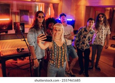 Portrait of multicultural small group of musicians standing at music recording studio and taking selfies. Blonde artist in foreground with cellphone taking selfie with her band members at music studio - Powered by Shutterstock