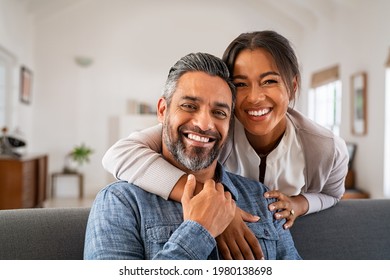 Portrait Of Multiethnic Couple Embracing And Looking At Camera Sitting On Sofa. Smiling African American Woman Hugging Mid Adult Man Sitting On Couch From Behind At Home. Happy Mixed Race Couple Laugh