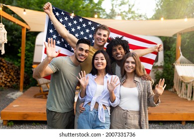 Portrait of multiethnic millennial friends with American flag waving at camera and smiling, posing near RV at camping site. Group of diverse young people celebrating patriotic holiday together outdoor - Powered by Shutterstock