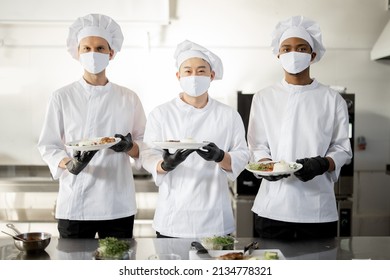 Portrait Of Multiracial Team Of Cooks Standing With Ready Meals For A Restaurant In The Kitchen. Chefs Wearing Uniform And Face Mask During Pandemic. Asian Chef With Latin And European Guys