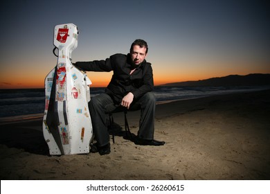 Portrait Of A Musician With Travel Cello Or Guitar Case, Sitting On Beach At Night. Shallow DOF.