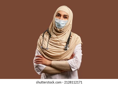Portrait of Muslim doctor woman wearing white robe and protective mask, arms crossed, standing near brown background. The concept of medicine, care, health.