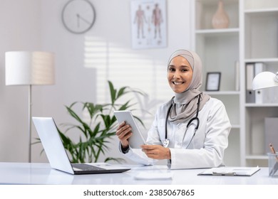 Portrait of Muslim female doctor in hijab, female doctor with tablet computer looking at camera with smile, working inside medical office, wearing white medical coat, sitting at table with laptop. - Powered by Shutterstock