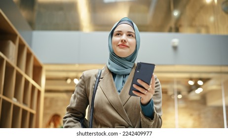 Portrait of Muslim Hijabi Woman Scrolling on her Smartphone While Walking in Office Hall. A happy Female Employee Finishing an Important Task and Smiling Arriving at Work. Low and Tilted Angle - Powered by Shutterstock