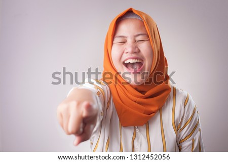 Portrait of muslim lady wearing hijab laughing hard and pointing forward, bully expression, close up head shot with selective focus