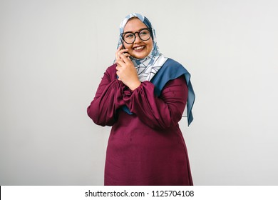 Portrait Of A Muslim Malay Woman Talking On A Smartphone. She Is Against A Plain White Background (studio) And Is Fashionably Dressed In A Colorful Tudung Head Scarf And Traditional Orange Garb. 