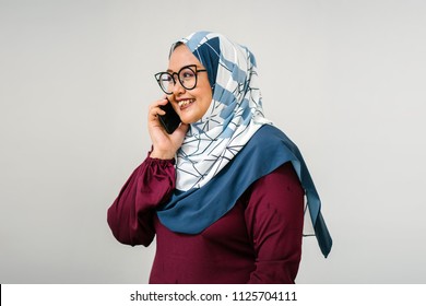 Portrait Of A Muslim Malay Woman Talking On A Smartphone. She Is Against A Plain White Background (studio) And Is Fashionably Dressed In A Colorful Tudung Head Scarf And Traditional Orange Garb. 